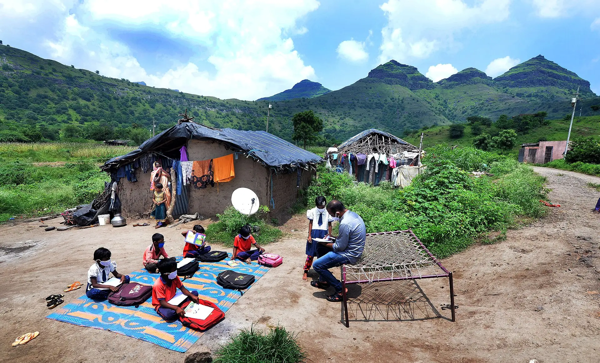 A teacher leads an open-air class for village children beside their home, with green hills behind